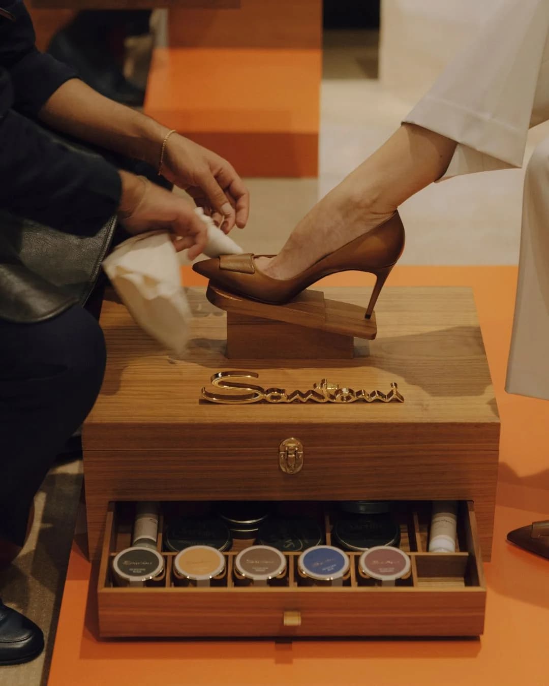 Santoni shoeshine service polishing a brown leather pump over a wooden care case