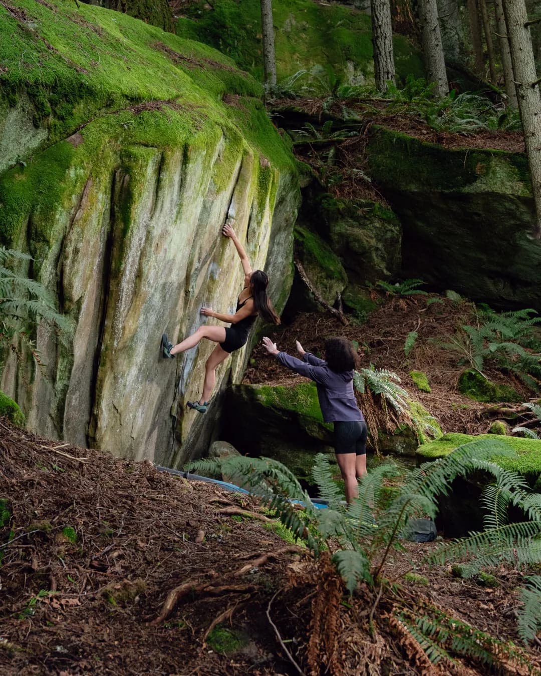 Woman bouldering on a moss-covered rock while a spotter stands below