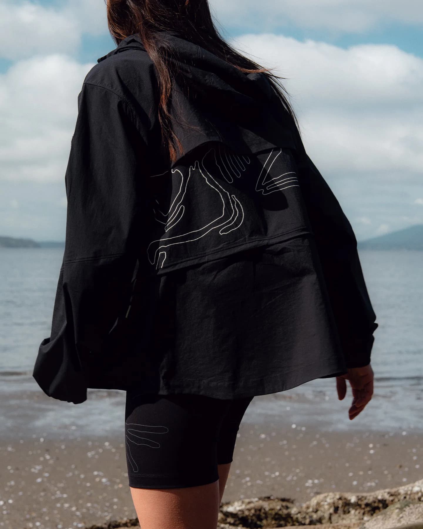 Model wearing a black Arc’teryx System_A jacket with white contour graphics by the water