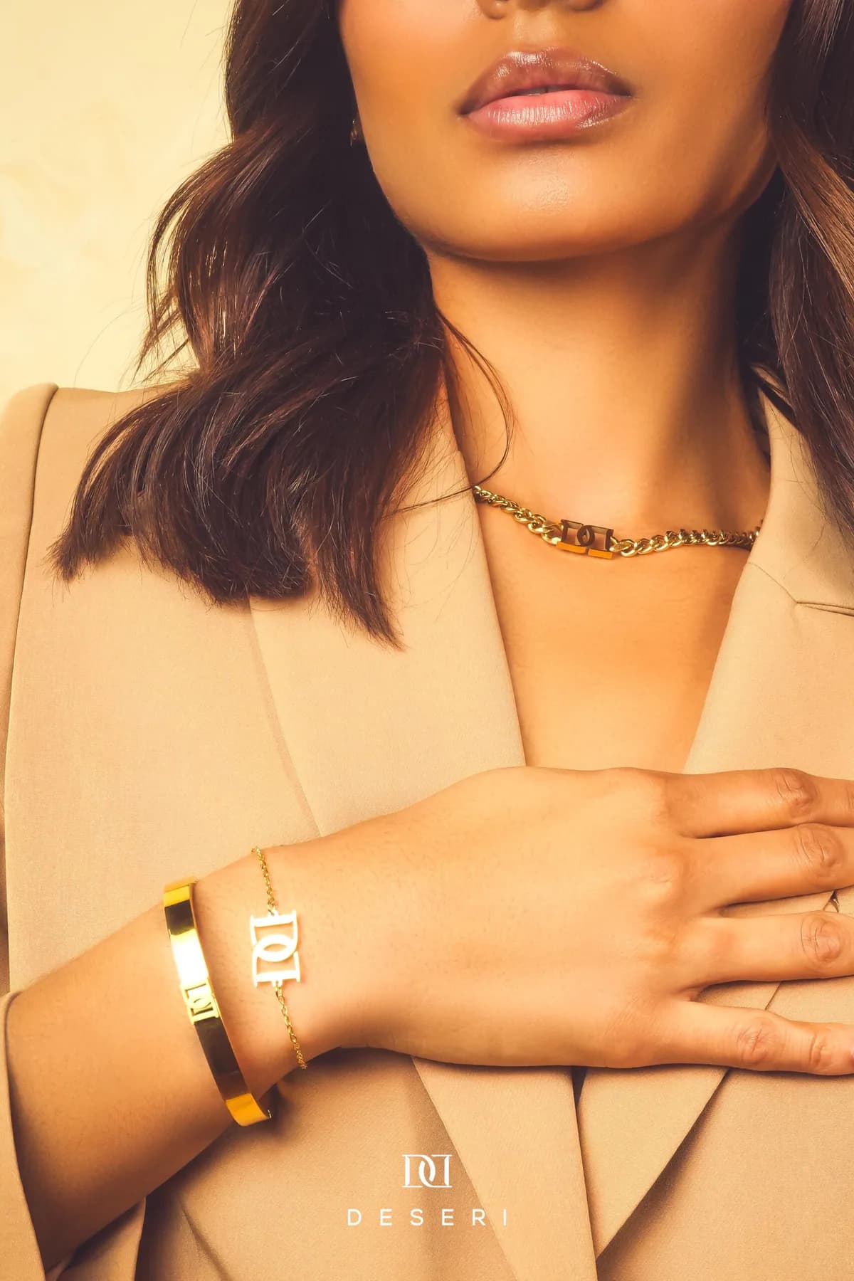 Model in a tan blazer wearing gold DESERI monogram jewelry