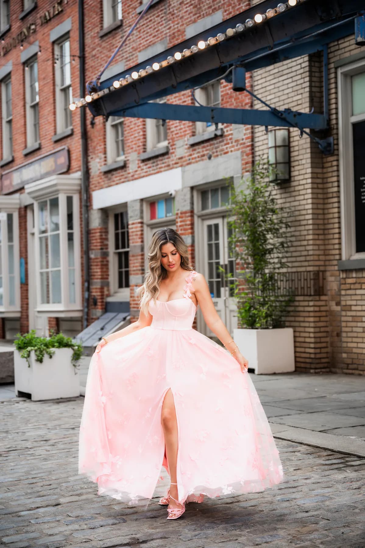 Model in a pale pink floral-appliqué gown standing on a cobblestone street
