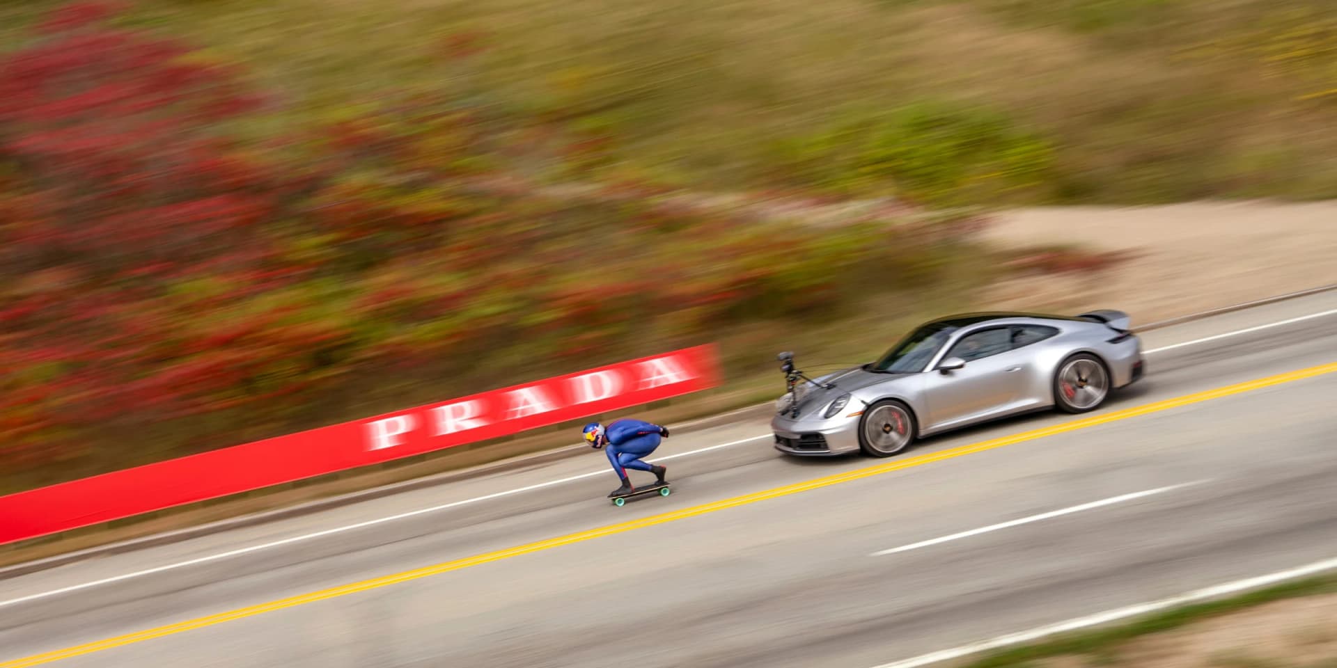Diego Poncelet sets off downhill on his skateboard on a closed Québec road, with a Porsche following behind.