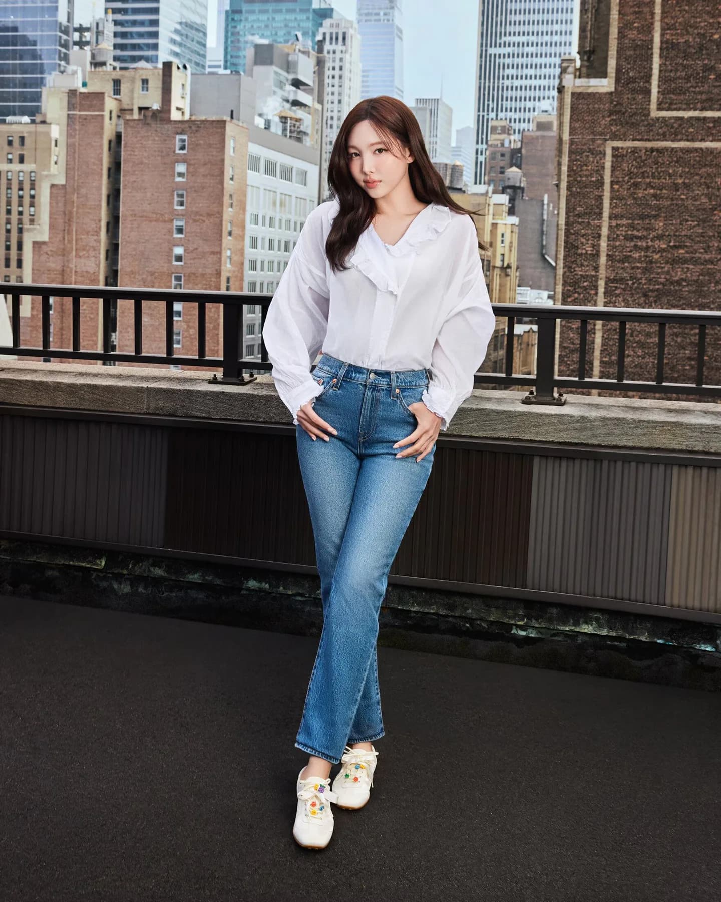 Nayeon wearing a white blouse, blue jeans, and white flats on a New York rooftop