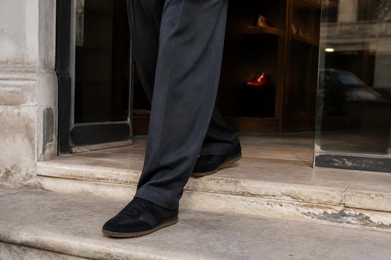 Person in charcoal trousers stepping outside in slim black archival-style sneakers