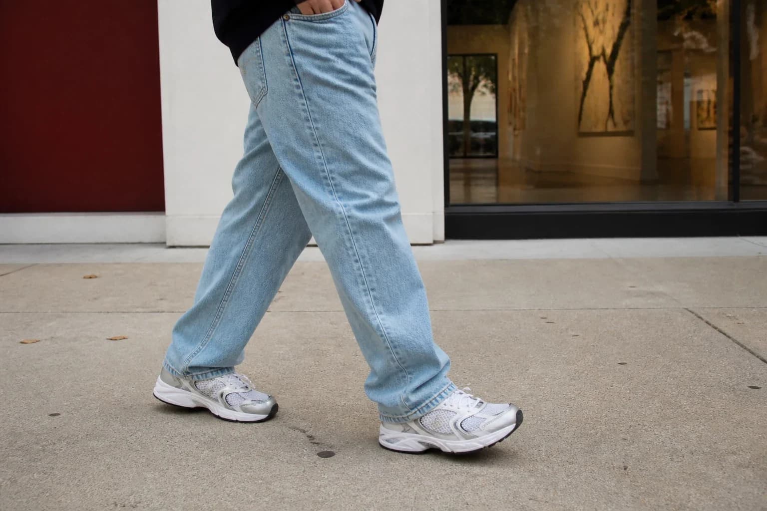 Person in relaxed light-wash jeans walking in silver mesh retro runners past a gallery