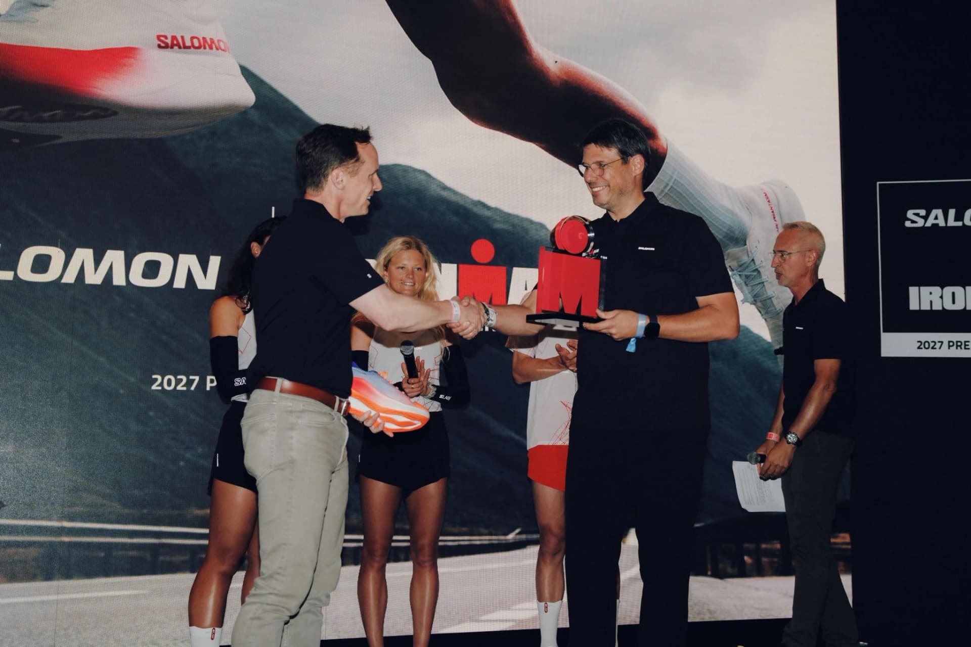 Two representatives shake hands beside a red IRONMAN emblem on stage.