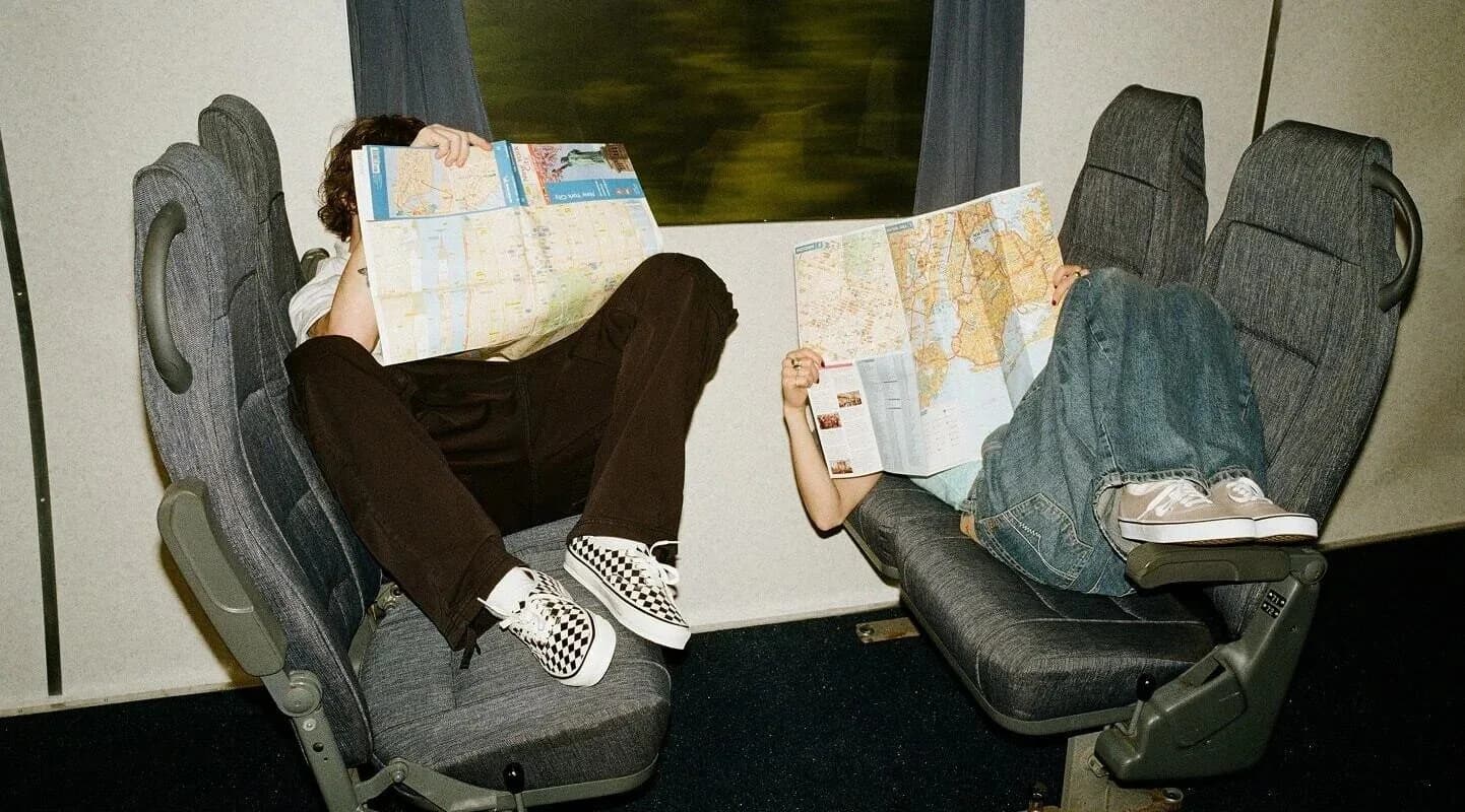 Two travelers wearing Vans sneakers hold maps over their faces on a train